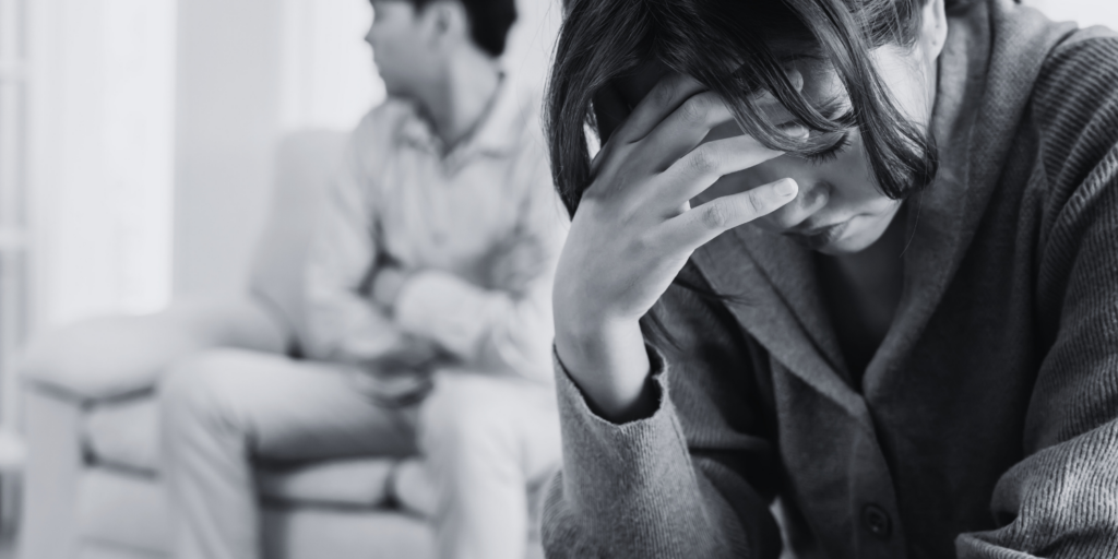 A woman placing her hand on her forehead and looking worried. Her partner a bit blurred in the background.