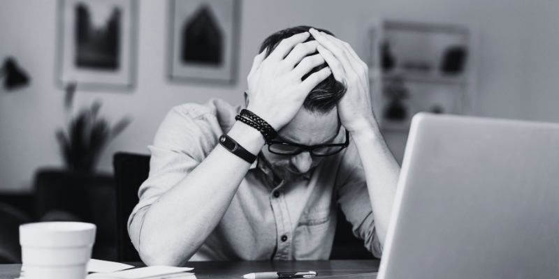 A man sitting in front of his laptop, resting his hands on his forehead, looking worried.